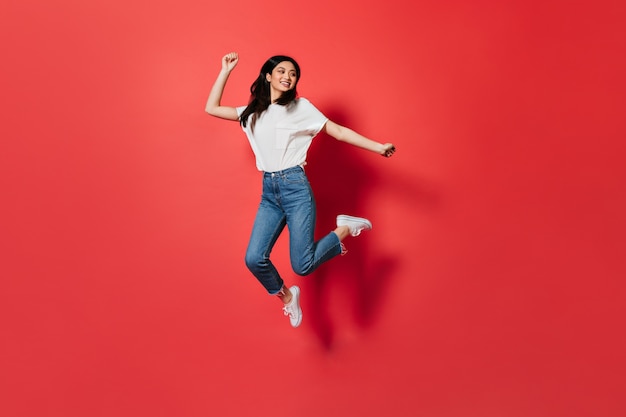 Mujer traviesa en camiseta blanca y jeans saltando sobre pared roja