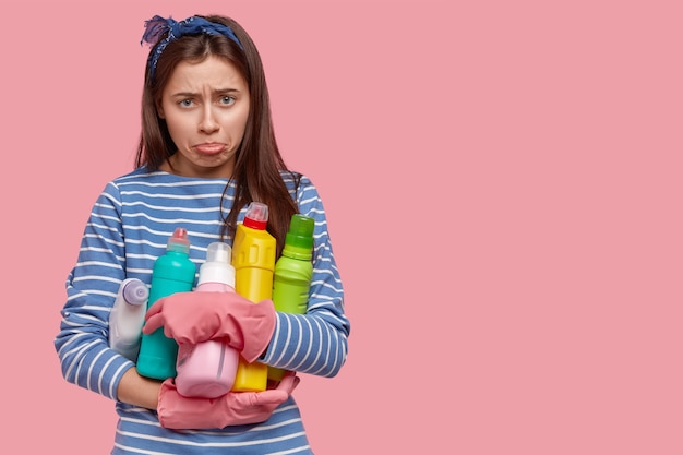 Una mujer triste y abatida se frunce el labio inferior, viste un jersey de marinero azul y guantes rosas, sostiene botellas de detergentes químicos, tiene mal humor y limpia en lugar de ir de fiesta. Espacio en blanco a un lado