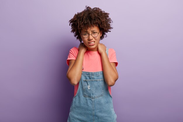 La mujer triste y cansada disgustada tiene el pelo afro, mantiene ambas manos en el cuello, sonríe de dolor, está cansada de trabajar en la computadora