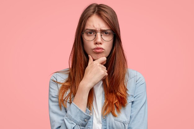 Mujer triste y descontenta con cara pecosa, sostiene la barbilla y frunce el ceño, está de mal humor, hace muecas de disgusto, usa chaqueta de mezclilla, aislada sobre fondo rosa