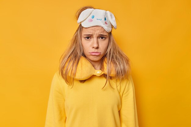 mujer triste por despertarse temprano en la mañana usa antifaz en la frente almohada para el cuello el suéter casual necesita más descanso aislado en amarillo. Gente durmiendo y durmiendo concepto.