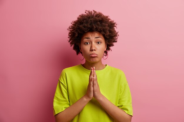 La mujer triste y esperanzada junta las palmas de las manos, suplica o pide disculpas, necesita tu ayuda, usa camiseta verde, posa contra la pared rosa. Por favor, hazme un favor por última vez. Concepto de lenguaje corporal.