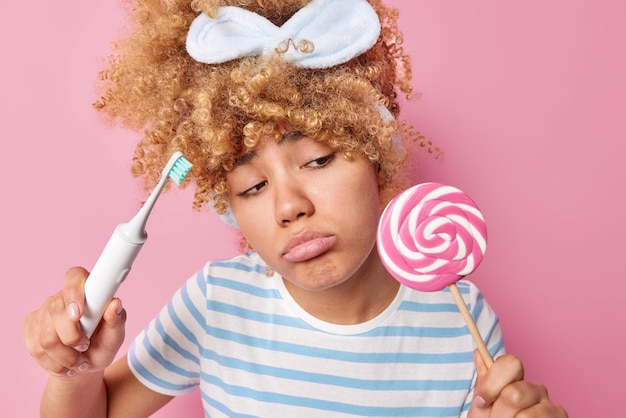 Una mujer triste y frustrada con el pelo rizado mira infelizmente a la piruleta que quiere comer dulce pero no puede, ya que tiene un efecto nocivo en los dientes, sostiene un cepillo de dientes eléctrico vestido de manera casual aislado en un fondo rosa