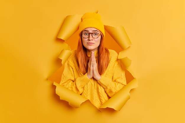 Una mujer triste sin gente tiene una expresión suplicante que mantiene las palmas juntas, pide disculpas, usa un suéter y un sombrero amarillo.