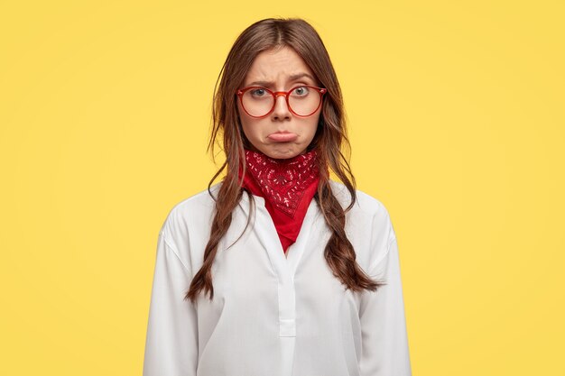 Mujer triste insultada frunce el labio inferior, molesta por noticias terribles, tiene dos trenzas ligeramente peinadas, usa gafas ópticas y camisa blanca, expresa emociones negativas, modelos sobre pared amarilla