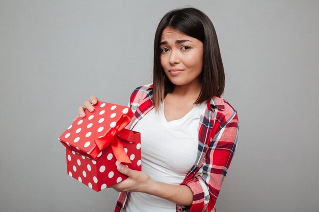 Mujer triste joven que sostiene el regalo sobre la pared gris.