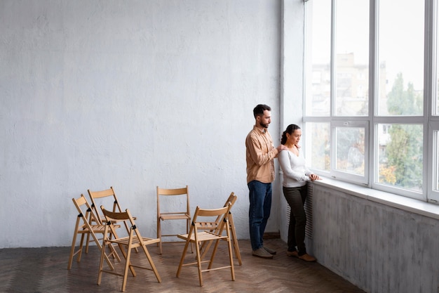 Mujer triste mirando por la ventana en una sesión de terapia de grupo con el hombre consolándola