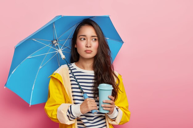 La mujer triste se siente deprimida durante un día lluvioso nublado, tiene depresión estacional, posa bajo un paraguas impermeable, usa un suéter de rayas y un impermeable