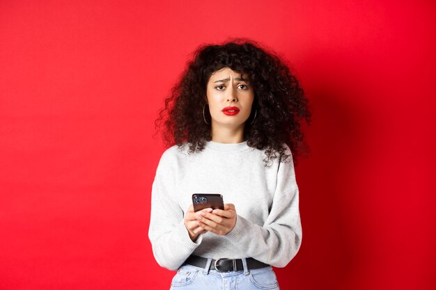 Mujer triste y sombría con cabello rizado, frunciendo el ceño y se siente molesta después de leer el mensaje del teléfono inteligente, de pie decepcionada contra el fondo rojo.