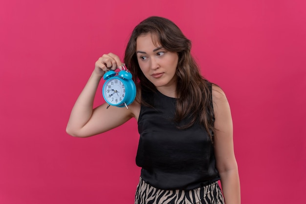 Mujer triste vestida con camiseta negra sosteniendo un despertador y escuchar en la pared rosa