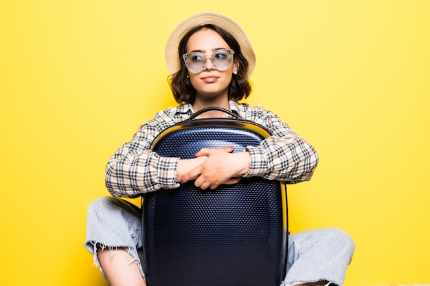 Mujer turista feliz en ropa casual de verano, sombrero sentado con maleta, mirando a un lado aislado en la pared naranja amarillo. Chica viajando al extranjero para viajar durante una escapada de fin de semana. Concepto de vuelo aéreo