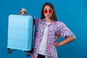 Foto gratuita mujer turista con gafas de sol rojas sosteniendo la maleta de viaje mirando confiados en azul aislado