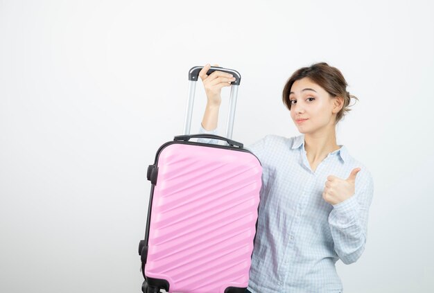 Mujer turista mostrando el pulgar hacia arriba y sosteniendo una maleta de viaje rosa. foto de alta calidad