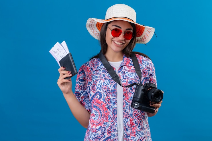 Foto gratuito mujer turista con sombrero con gafas de sol rojas de pie con cámara fotográfica sosteniendo pasaporte y boletos sonriendo alegremente, listo para vacaciones en azul aislado