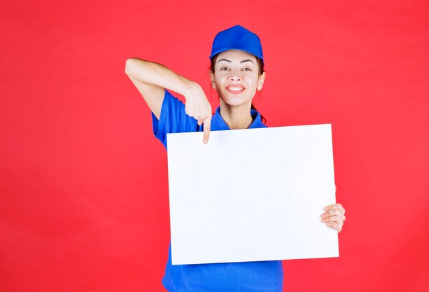 Mujer en uniforme azul y boina sosteniendo un mostrador de información cuadrado blanco y sintiéndose positivo.