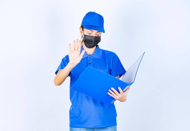 Mujer en uniforme azul y mascarilla negra sosteniendo una carpeta azul y deteniendo algo.