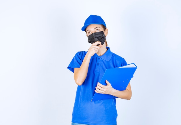 Mujer en uniforme azul y mascarilla negra sosteniendo una carpeta azul y parece confundida y pensativa.