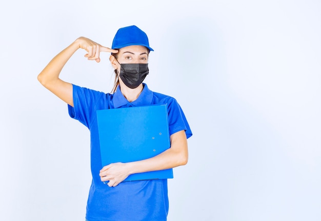 Mujer en uniforme azul y mascarilla negra sosteniendo una carpeta azul y parece confundida y pensativa.