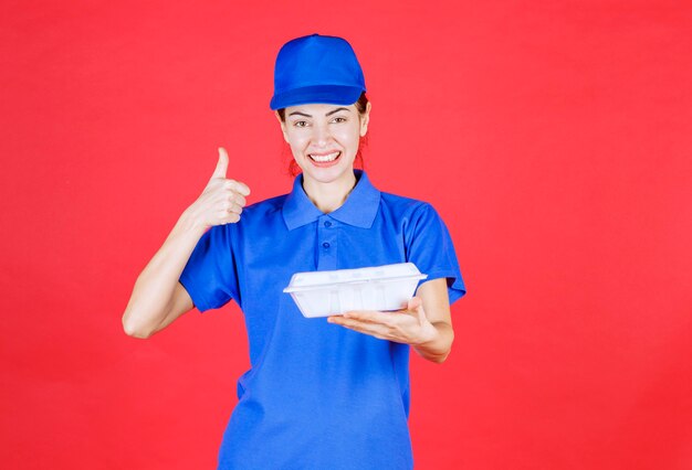 Mujer en uniforme azul sosteniendo una caja de comida para llevar de plástico blanco para la entrega y mostrando el signo de satisfacción.