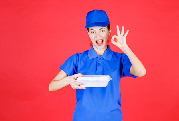 Mujer en uniforme azul sosteniendo una caja de comida para llevar de plástico blanco para la entrega y mostrando el signo de satisfacción.