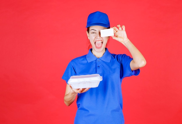 Mujer en uniforme azul sosteniendo una caja de comida para llevar de plástico blanco para la entrega y presentando su tarjeta de visita al cliente.