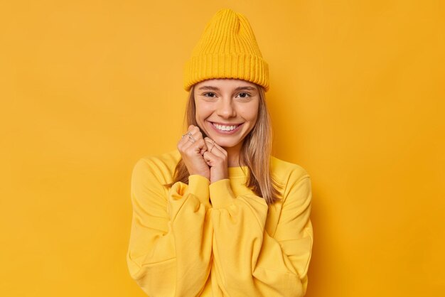 La mujer usa un jersey casual y un sombrero mantiene las manos cerca de la cara y sonríe agradablemente aislado en amarillo. Concepto de emociones y expresiones de rostro humano