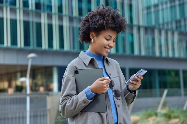 Mujer usa tecnologías modernas en la ciudad sostiene un teléfono móvil y una tableta digital portátil sonríe alegremente mientras lee un mensaje de texto agradable y usa ropa elegante