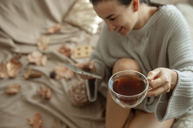 Una mujer usa el teléfono y sostiene una taza de té mientras está sentada en la cama con un acogedor suéter de punto, fondo borroso.