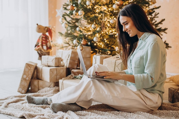 Mujer usando laptop y sentada junto al árbol de Navidad