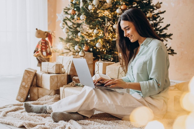 Mujer usando laptop y sentada junto al árbol de Navidad