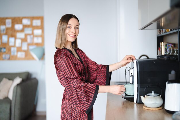 Mujer usando una máquina de café para hacer una gran taza de café en casa Mujer vistiendo una bata de seda en casa mientras prepara un café con leche