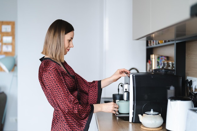 Mujer usando una máquina de café para hacer una gran taza de café en casa Mujer vistiendo una bata de seda en casa mientras prepara un café con leche