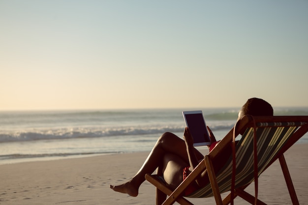 Mujer usando tableta digital mientras se relaja en una silla de playa en la playa