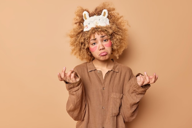 Una mujer vacilante y descontenta con el pelo rubio rizado y tupido aplica parches de belleza, se encoge de hombros y tiene una expresión de mal humor vestida con un traje de dormir que no sabe qué hacer aislada sobre un fondo marrón