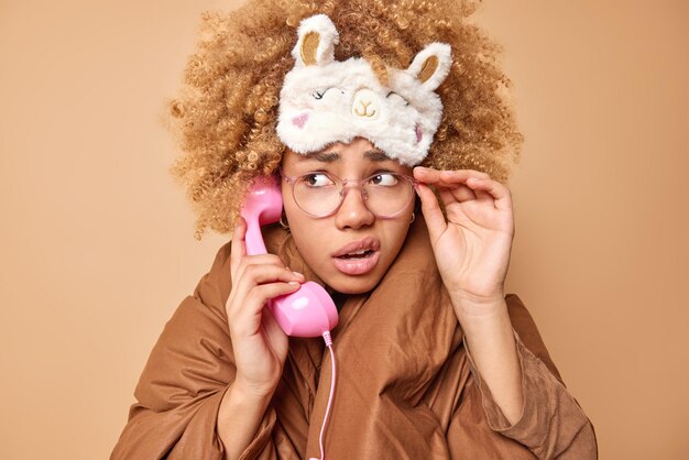 Mujer vacilante despistada parece avergonzada usa anteojos transparentes tiene conversación a través de un teléfono retro sostiene el auricular cerca de la oreja envuelto en una manta aislada sobre fondo beige habla antes de dormir