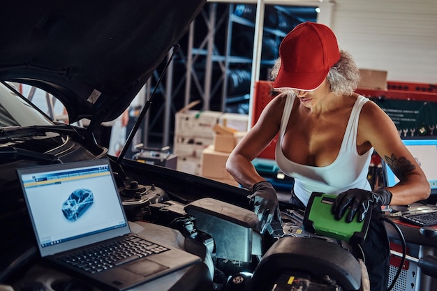 Una mujer varonil enfocada está haciendo diagnósticos de automóviles con ayuda de computadoras en el servicio de automóviles.