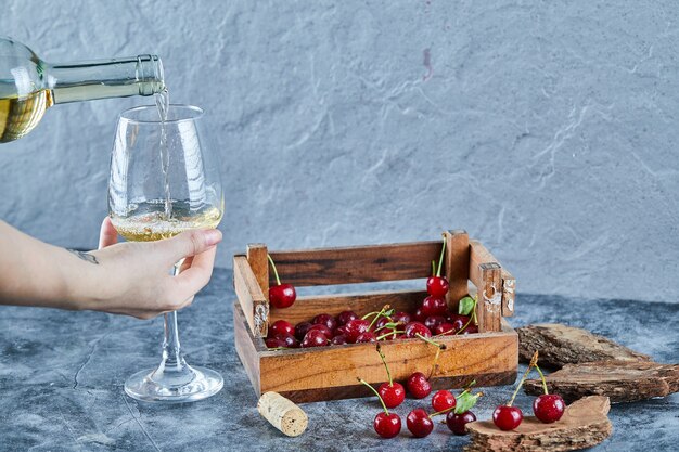 Mujer vertiendo vino blanco mientras sostiene un vaso y caja de madera de cerezas sobre superficie azul