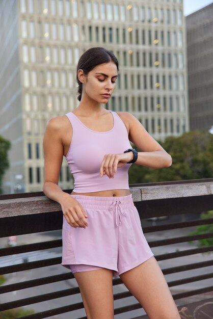 mujer vestida con camiseta sin mangas y pantalones cortos comprueba el tiempo en poses de smartwatch en el puente en el edificio de la ciudad moderna espera a alguien para comenzar a entrenar juntos. Rutina de fitness para la salud