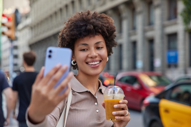 Mujer vestida casualmente bebidas batido toma selfie graba videos sonríe felizmente posa en una carretera muy transitada con transporte en la calle