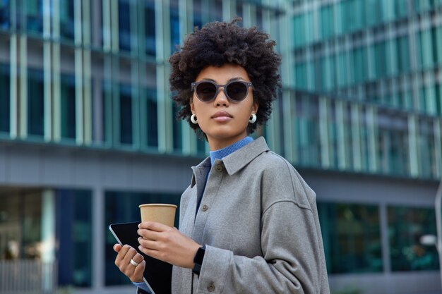 Mujer vestida con chaqueta gris bebe café de un vaso desechable de papel tiene poses de tableta moderna contra el edificio de negocios moderno en el exterior lleva gafas de sol mira hacia otro lado