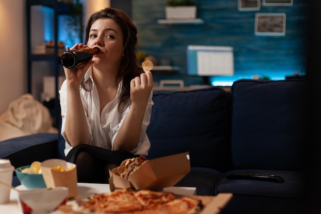 Mujer vestida informal bebiendo cerveza de botella mientras sostiene una papa frita mirando programa de comedia de comedia de televisión. Persona sentada en el sofá viendo la televisión frente a la mesa con comida rápida.