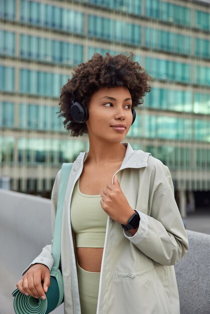 Mujer vestida con mallas recortadas y chaqueta lleva enrollada karemat escucha música motivacional a través de auriculares plantea contra edificio moderno