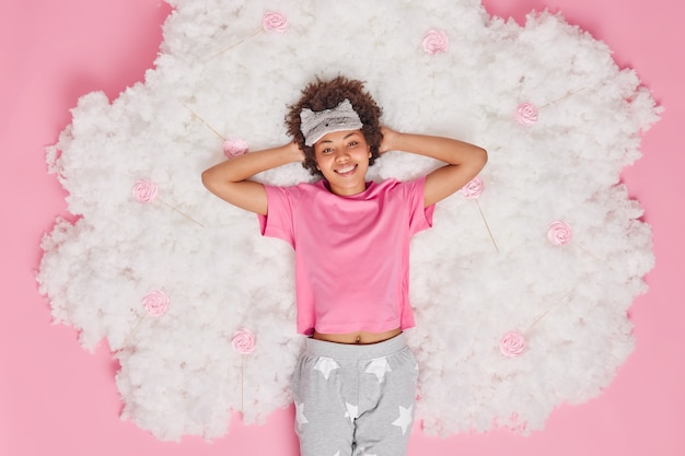 Foto gratuita mujer vestida con pijama mantiene las manos detrás de la cabeza sonríe alegremente se siente renovada después de ver sueños agradables y poses para dormir en la nube blanca