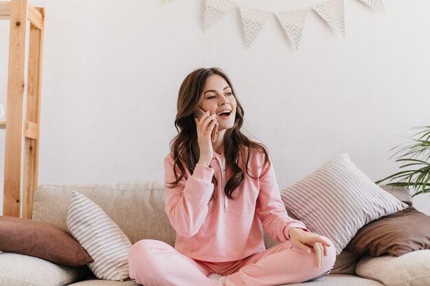 Mujer vestida con pijama rosa sentada en su sofá rodeada de suaves almohadas y hablando por teléfono