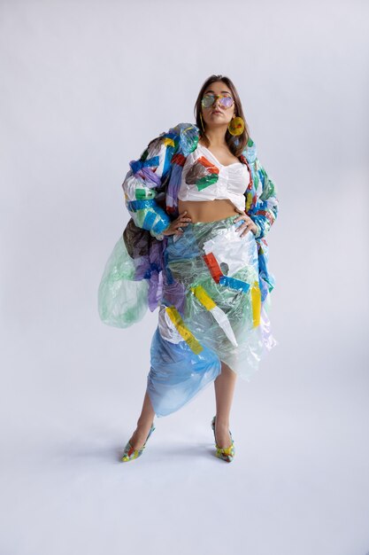 Mujer vestida de plástico en la pared blanca. Modelo femenino en ropa y zapatos hechos de basura. Moda, estilo, reciclaje, concepto ecológico y medioambiental. Demasiada contaminación, la estamos comiendo y tomándola.