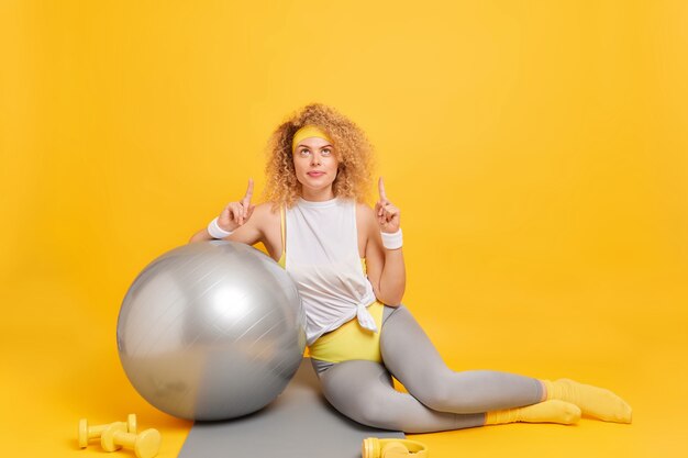 Mujer vestida con ropa deportiva, los puntos anteriores con los dedos índices se inclina sobre la bola de fitness demuestra el lugar para su anuncio en amarillo