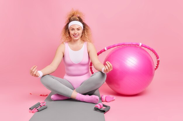 Mujer vestida con ropa deportiva se sienta en posición de loto rodeado de equipamiento deportivo se sienta en la estera de fitness