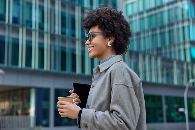 Mujer vestida con ropa de moda bebidas café para llevar tiene poses de tableta digital contra el edificio de la ciudad de cristal moderno paseos fuera durante el día