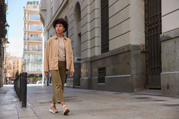 Mujer vestida con ropa de moda tiene un recorrido por la ciudad durante los fines de semana mira hacia otro lado camina en el centro de la ciudad sobre el pavimento cerca de los edificios antiguos llega al destino