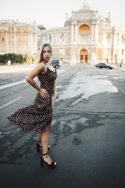 Mujer en vestido ajustado en los tacones altos está de pie en la carretera mojada en frente del antiguo edificio arquitectónico